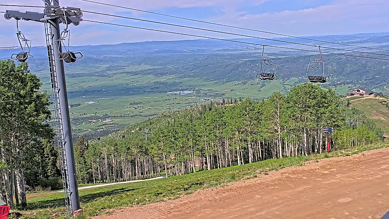 Two empty chairlift seats hang from cables above a mountain vista of green trees and a valley under a mostly sunny sky.