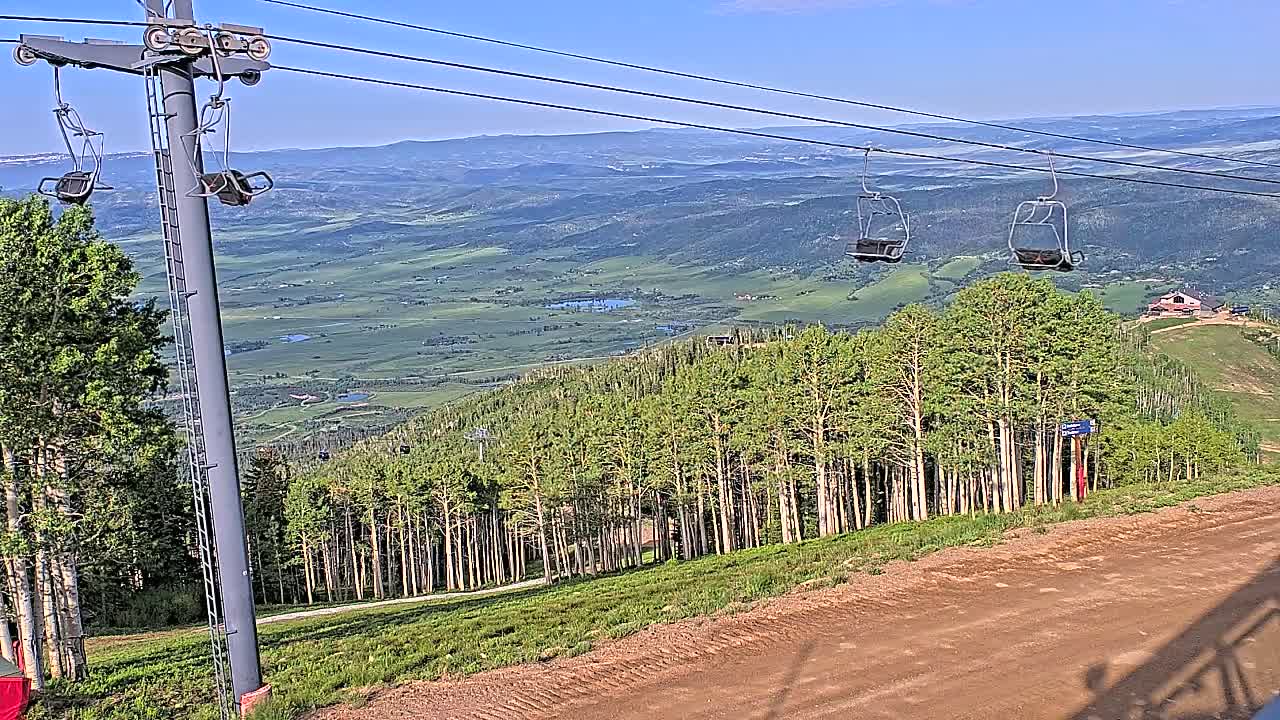 A chairlift ascends a mountain on a clear, sunny day, overlooking a vast valley with green fields and forests.
