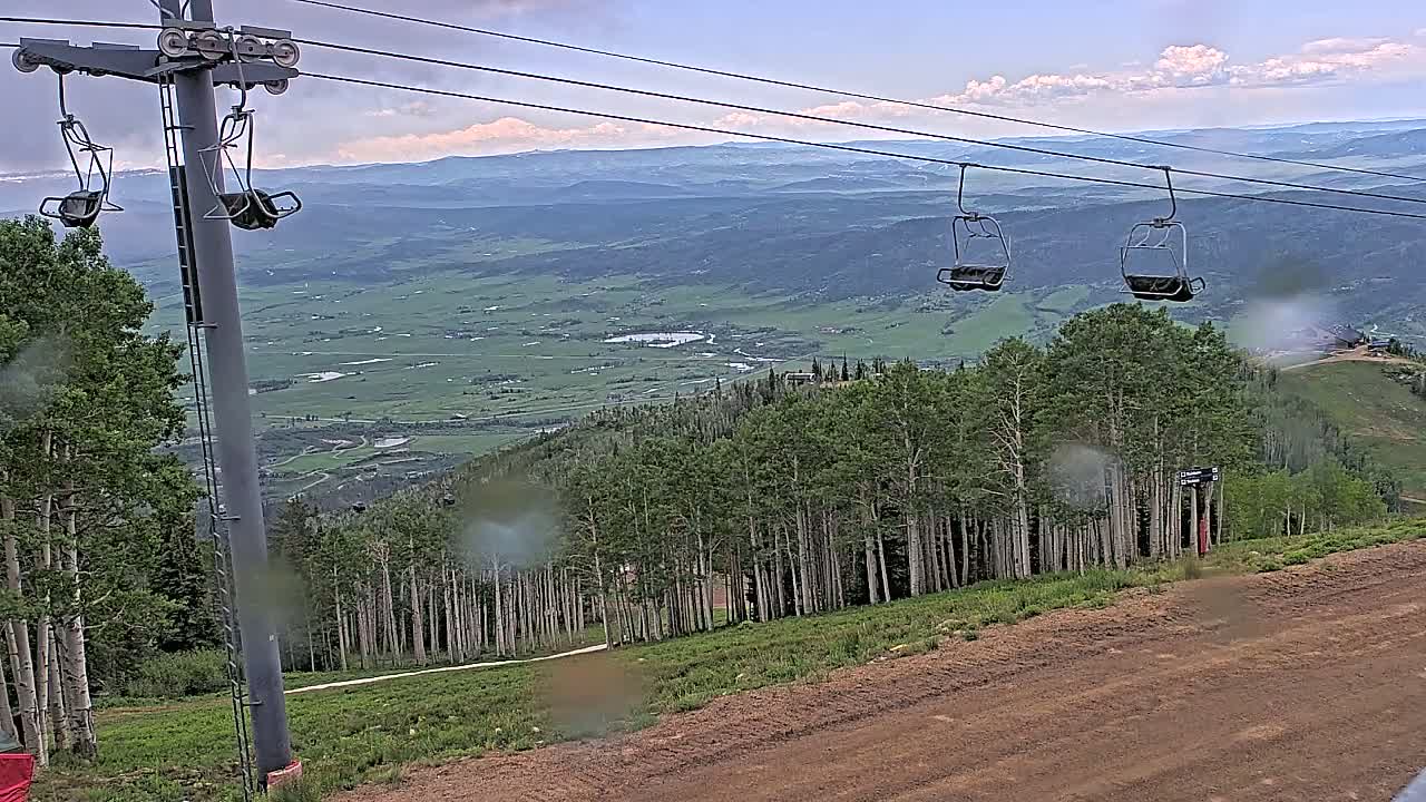 Two empty chairlifts hang from a cable against a backdrop of a mountainous valley under a partly cloudy sky, with rain obscuring parts of the view.