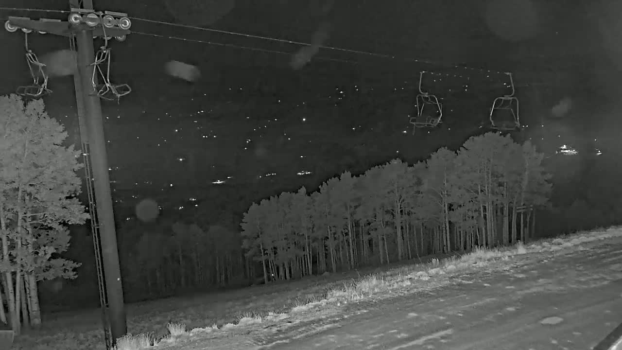 A nighttime, snowy scene shows a ski lift with empty chairs against a background of a distant town and a stand of trees.