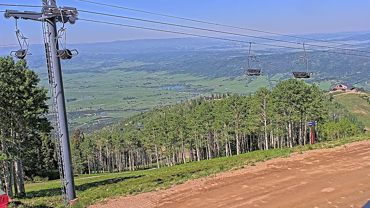 A chairlift ascends a mountain on a clear, sunny day, overlooking a vast valley dotted with greenery.