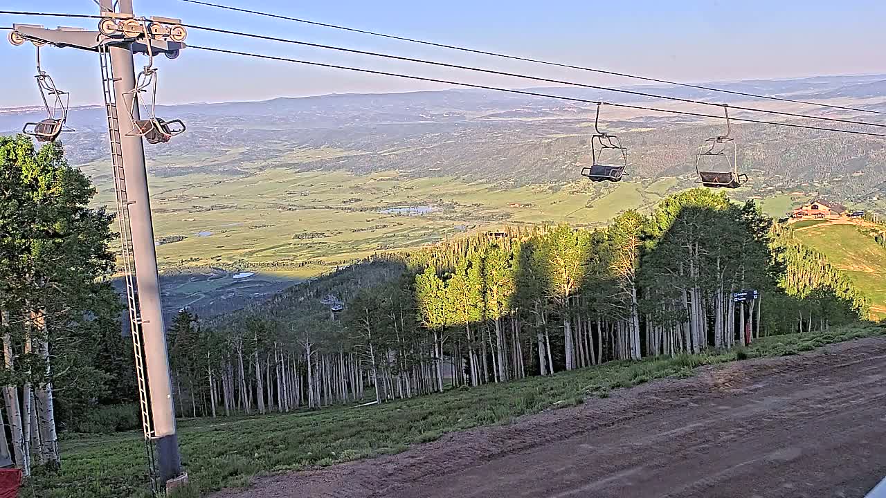 Two empty chairlifts ascend a mountainside overlooking a vast, sunlit valley under a clear sky.