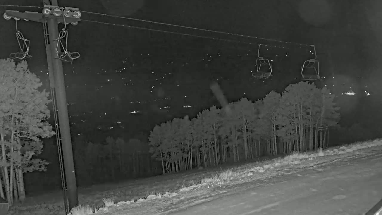 At night, a ski lift with empty chairs is visible against a backdrop of a snow-covered landscape, trees, and a distant town illuminated by lights.