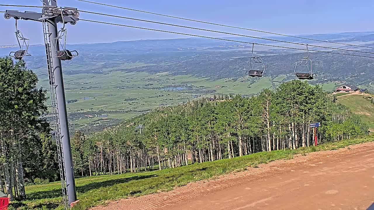A ski lift with empty chairs overlooks a vast, verdant valley under a clear, sunny sky.