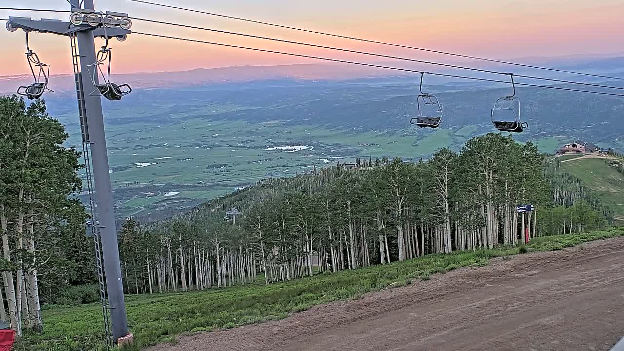 An empty chairlift ascends a mountain at sunset overlooking a vast, verdant valley.