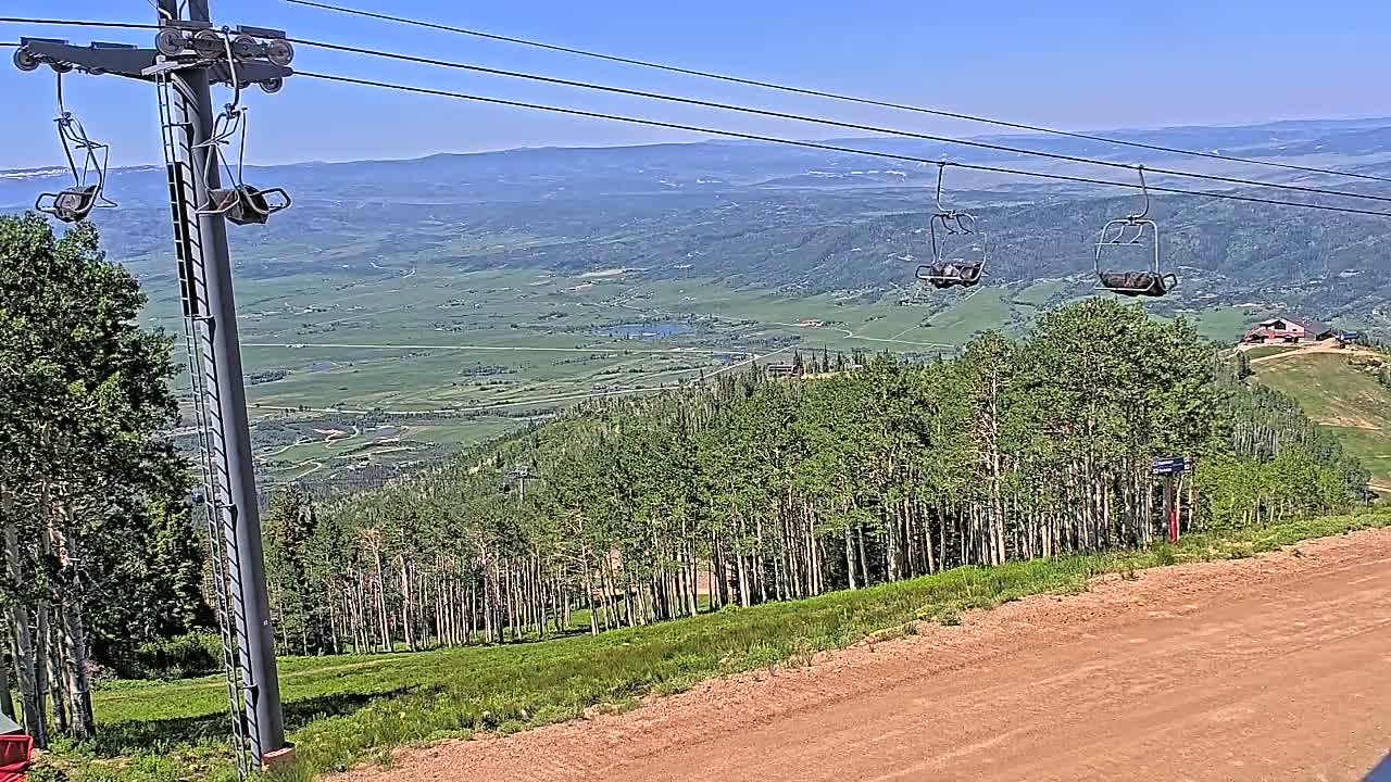 A chairlift ascends a mountain on a clear, sunny day, overlooking a vast valley with green fields and forests.