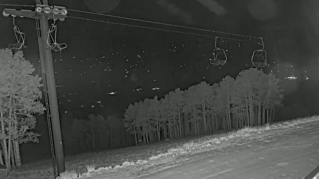 At night, empty ski lift chairs hang from cables above a snow-covered road and a stand of trees, with distant lights illuminating a valley below.