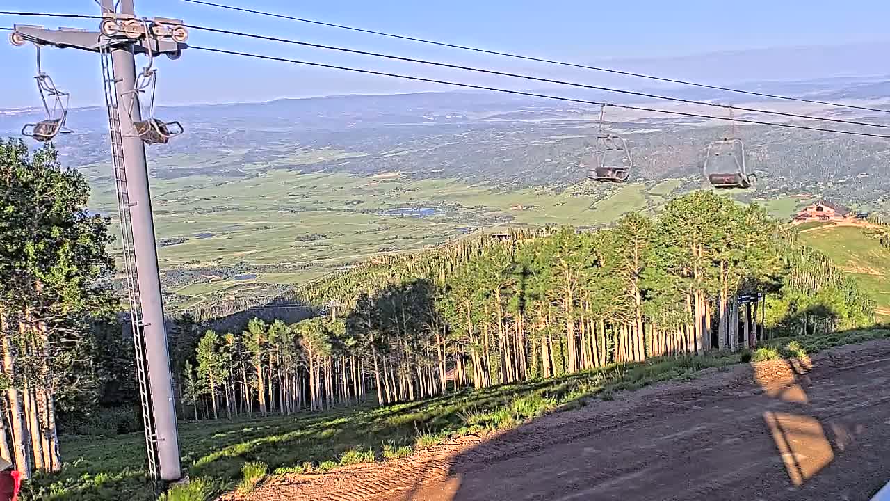 A chairlift ascends a mountain slope overlooking a vast green valley under a clear, sunny sky.