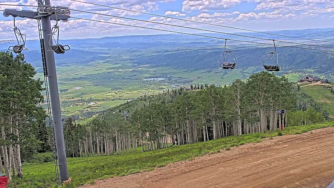 A mountaintop view on a partly cloudy day shows a chairlift, a dirt road, and a vast green valley below, dotted with trees and a few buildings.