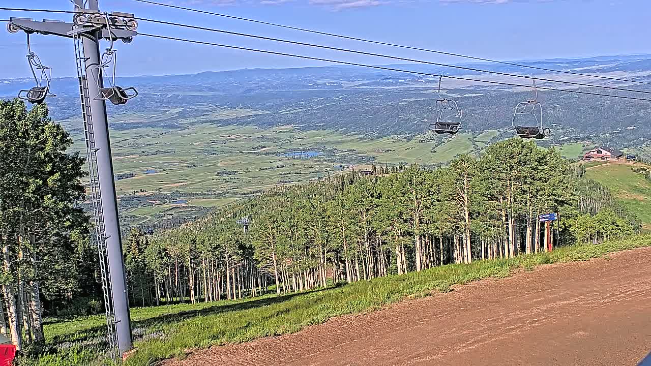 A chairlift ascends a mountainside on a clear, sunny day, overlooking a vast, verdant valley.
