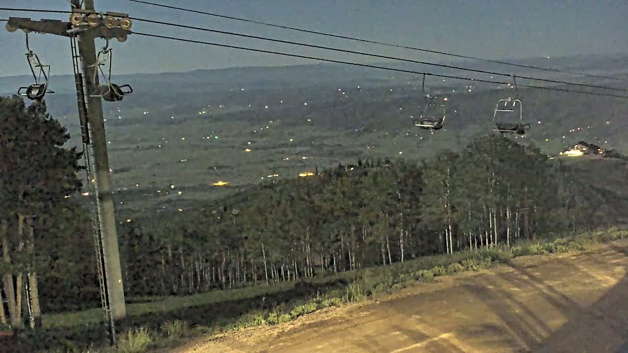 A chairlift hangs empty against a night sky overlooking a valley dotted with lights and a dark forest.