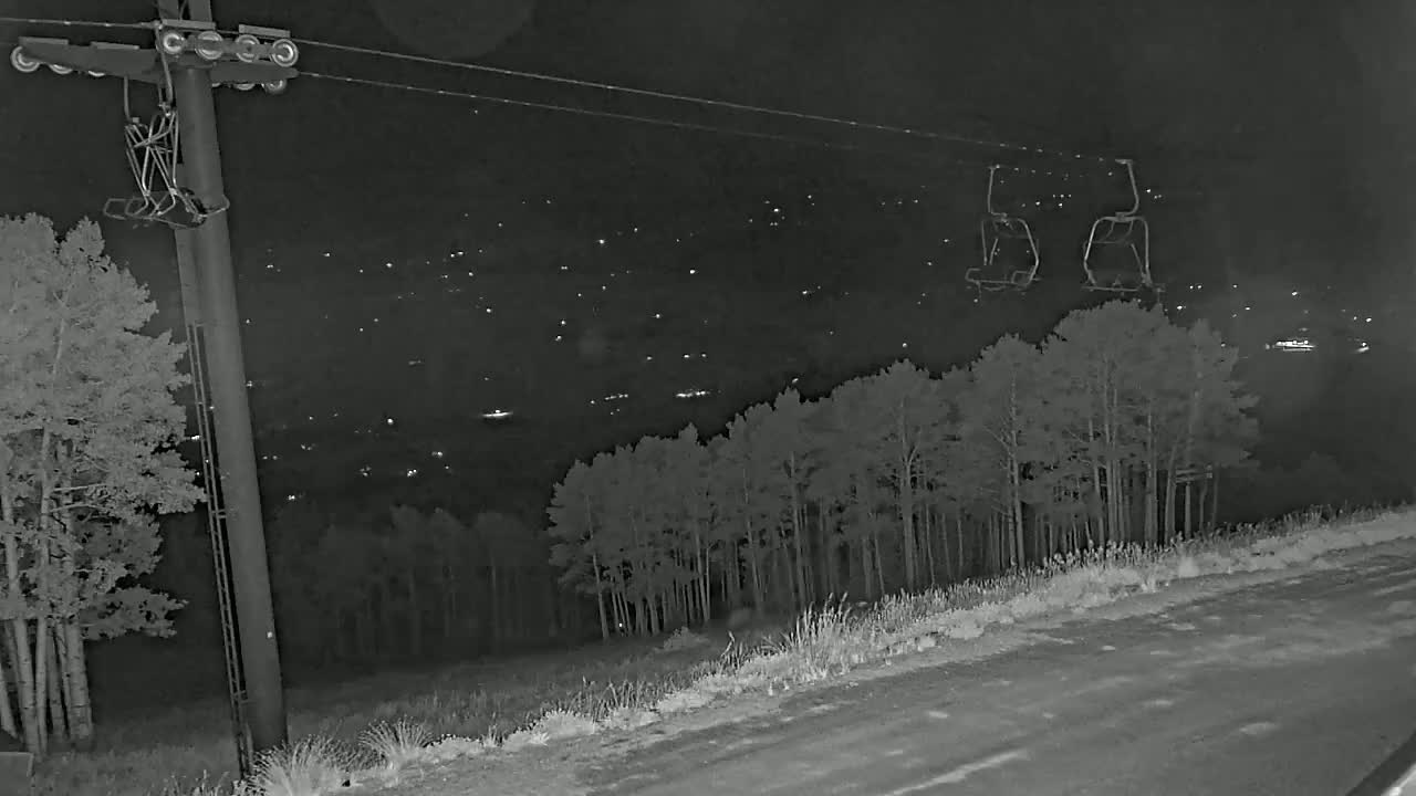 A nighttime view of a ski lift chair hanging from a cable above a dark forest and a distant town illuminated by lights.