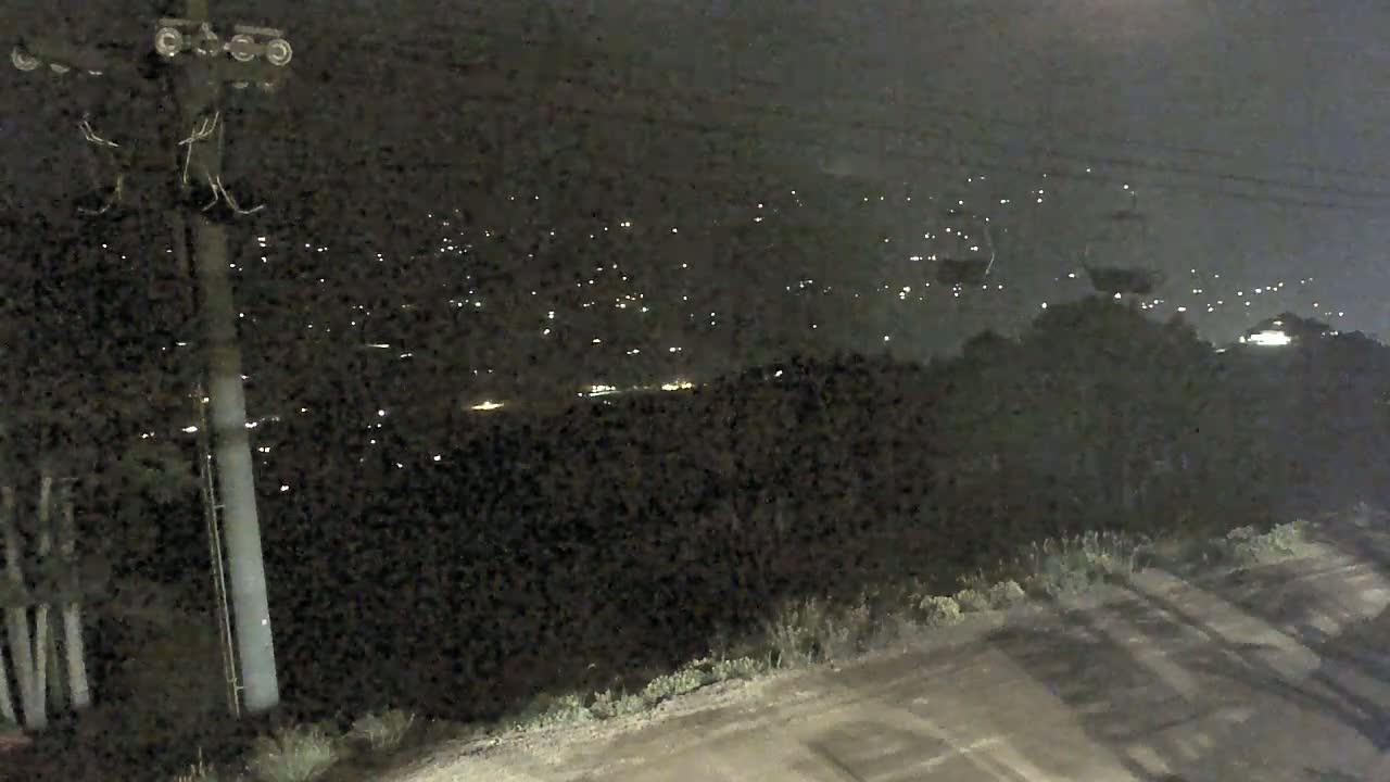 A nighttime view of a ski lift chair hanging from a cable above a dark forest and a distant town illuminated by lights.