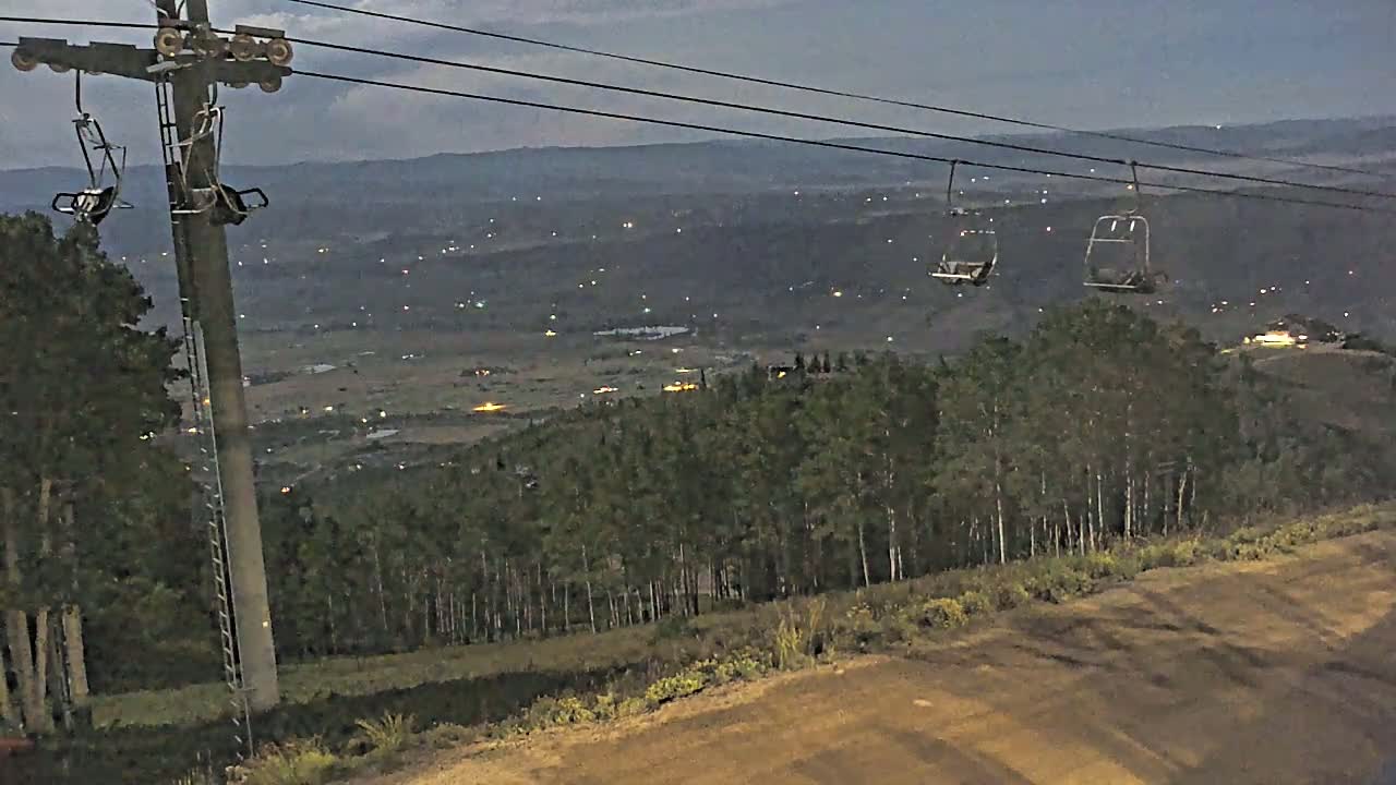Two empty chairlifts hang from cables above a dark, tree-covered hillside overlooking a valley speckled with distant lights under a twilight sky.