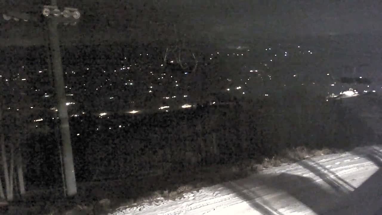 A clear night view reveals a snowy slope in the foreground, a ski lift tower to the left, and a distant town illuminated by numerous lights against a dark, mountainous landscape.