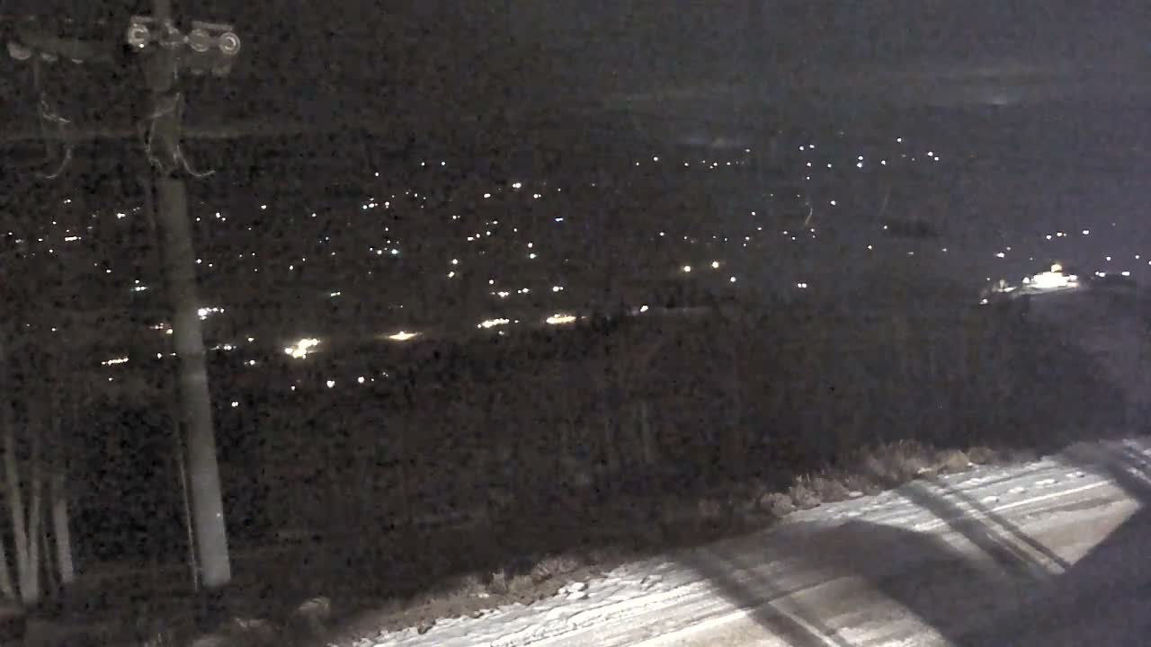 A dark, outdoor night scene displays distant city lights across a snowy landscape with a utility pole on the left, under clear, cold weather conditions.
