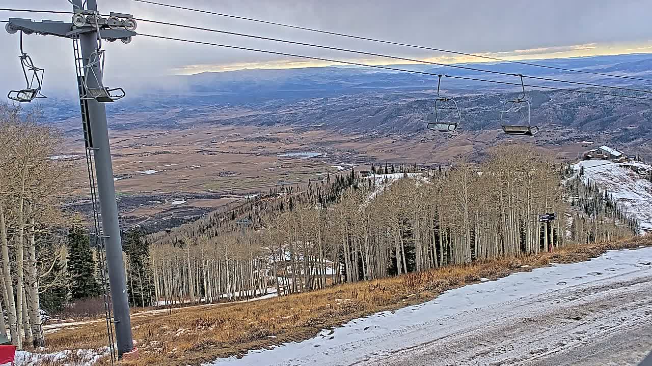 An overcast winter day presents a ski slope with patches of snow and bare trees, featuring a ski lift tower with empty chairs, all overlooking a vast valley and distant mountains under a cloudy sky.
