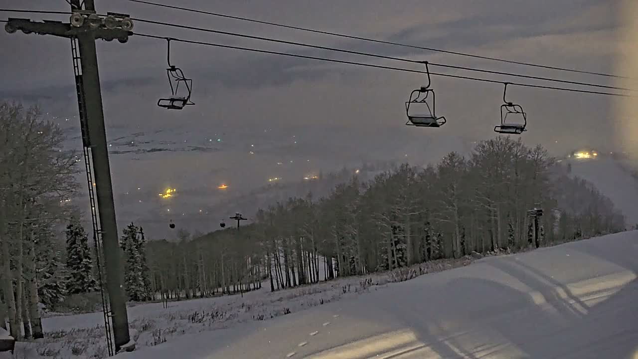 A snowy ski slope is visible at night under an overcast sky, featuring empty ski lift chairs suspended above snow-dusted trees and distant artificial lights.