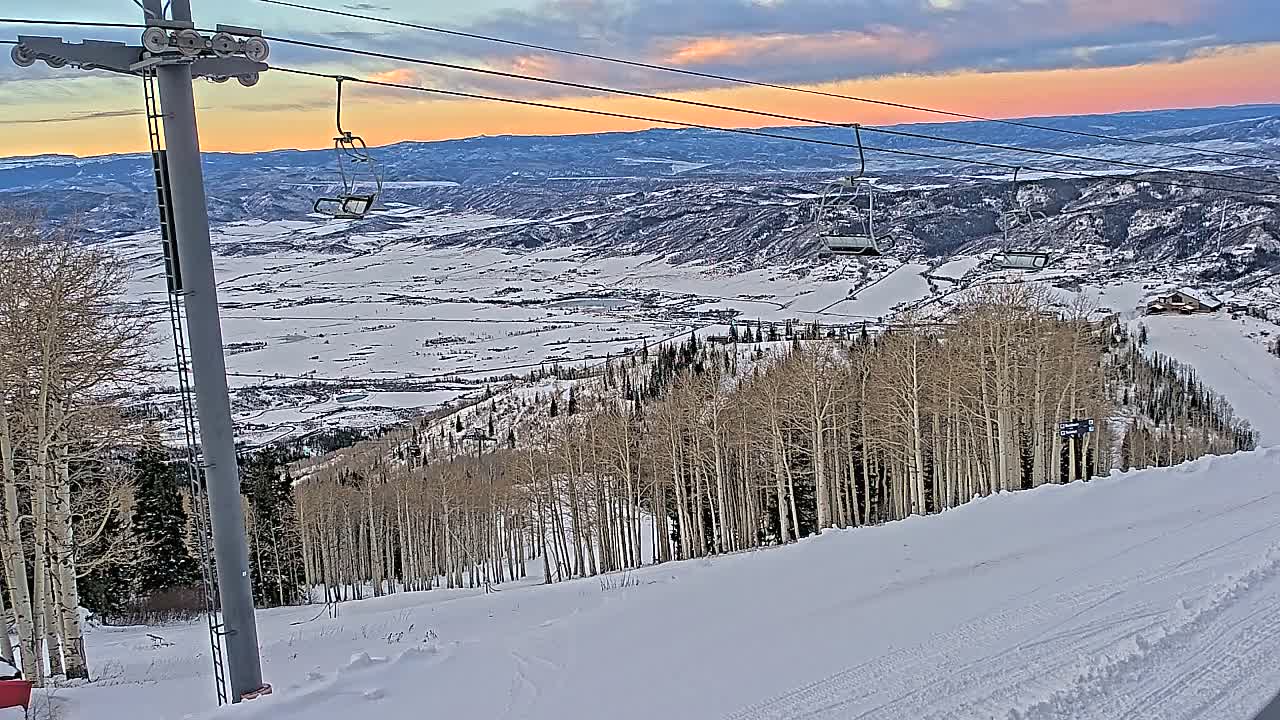 A snowy ski slope is visible at night under an overcast sky, featuring empty ski lift chairs suspended above snow-dusted trees and distant artificial lights.