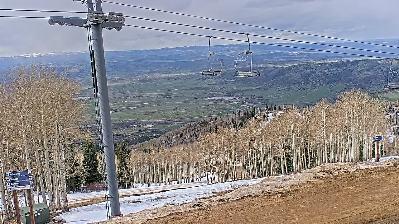 A snowy ski slope is visible at night under an overcast sky, featuring empty ski lift chairs suspended above snow-dusted trees and distant artificial lights.