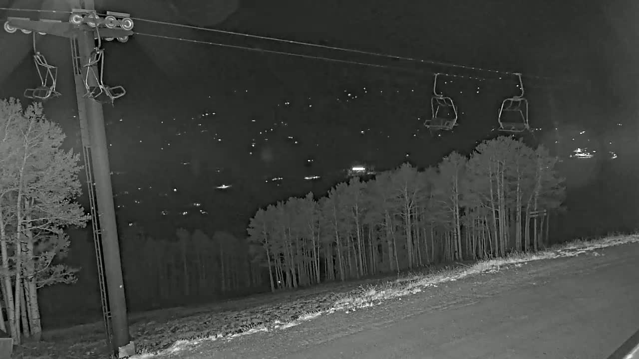 At night, a ski lift with empty chairs extends over a snow-covered slope and a distant town illuminated by lights.