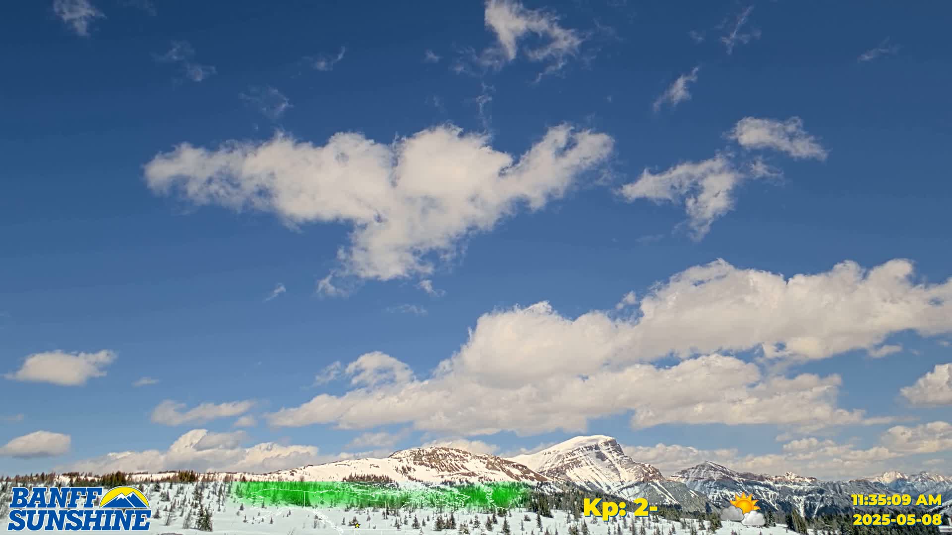 A mostly sunny sky with scattered cumulus clouds overlooks snow-covered mountains and evergreen trees.
