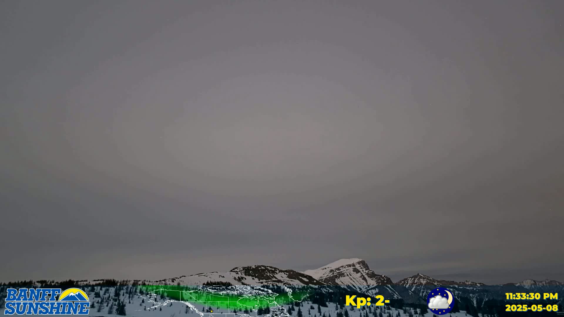 A mostly cloudy, overcast sky hangs over snow-covered mountains and a dark coniferous forest.