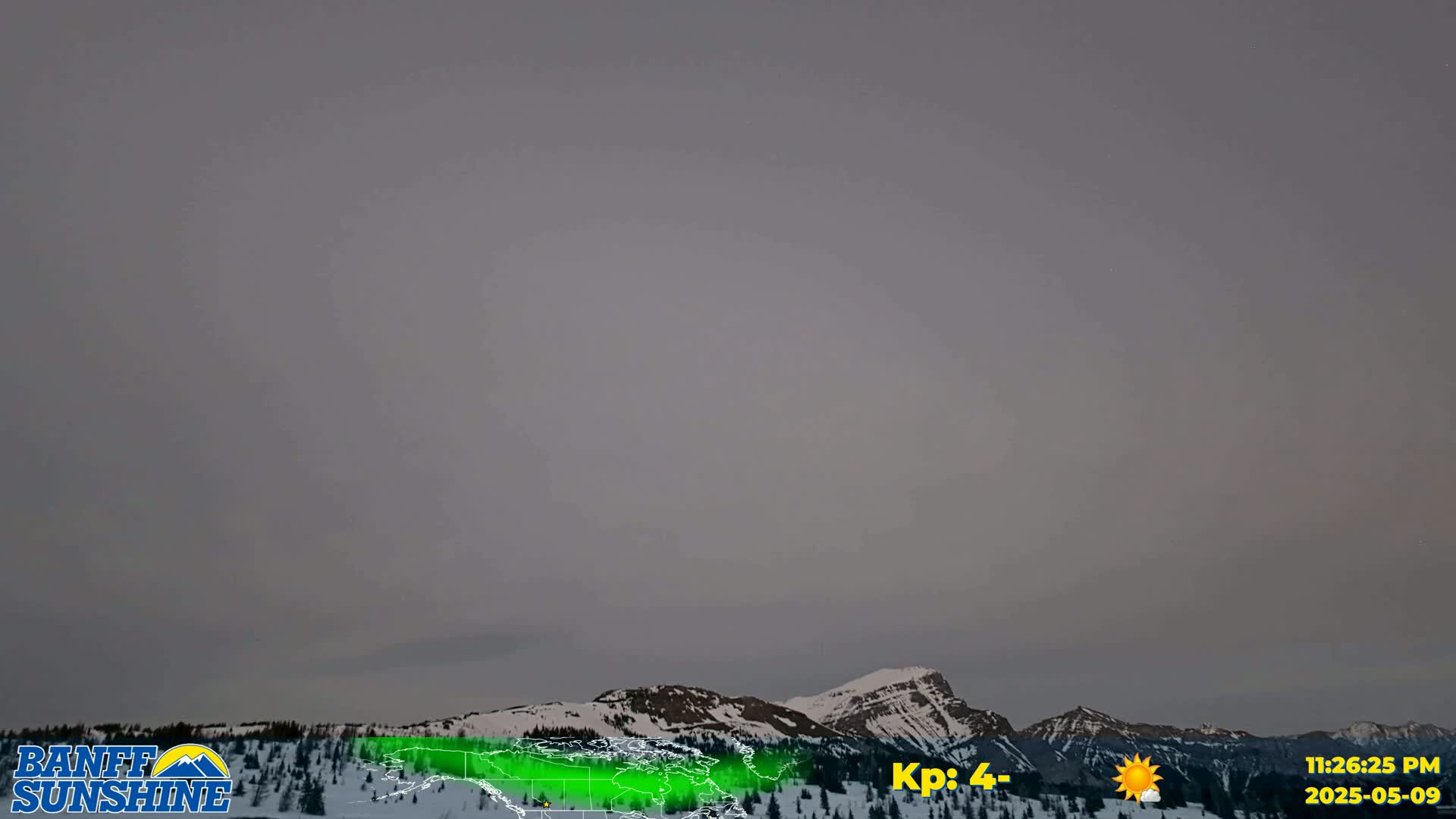 A faint aurora borealis glows over a snow-covered mountain range under an overcast sky.