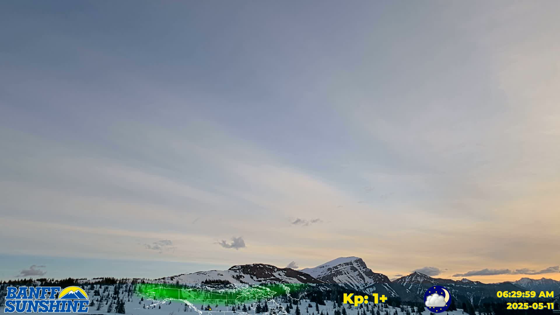 A sunrise illuminates wispy clouds over snow-covered mountains under a pale, clear sky.