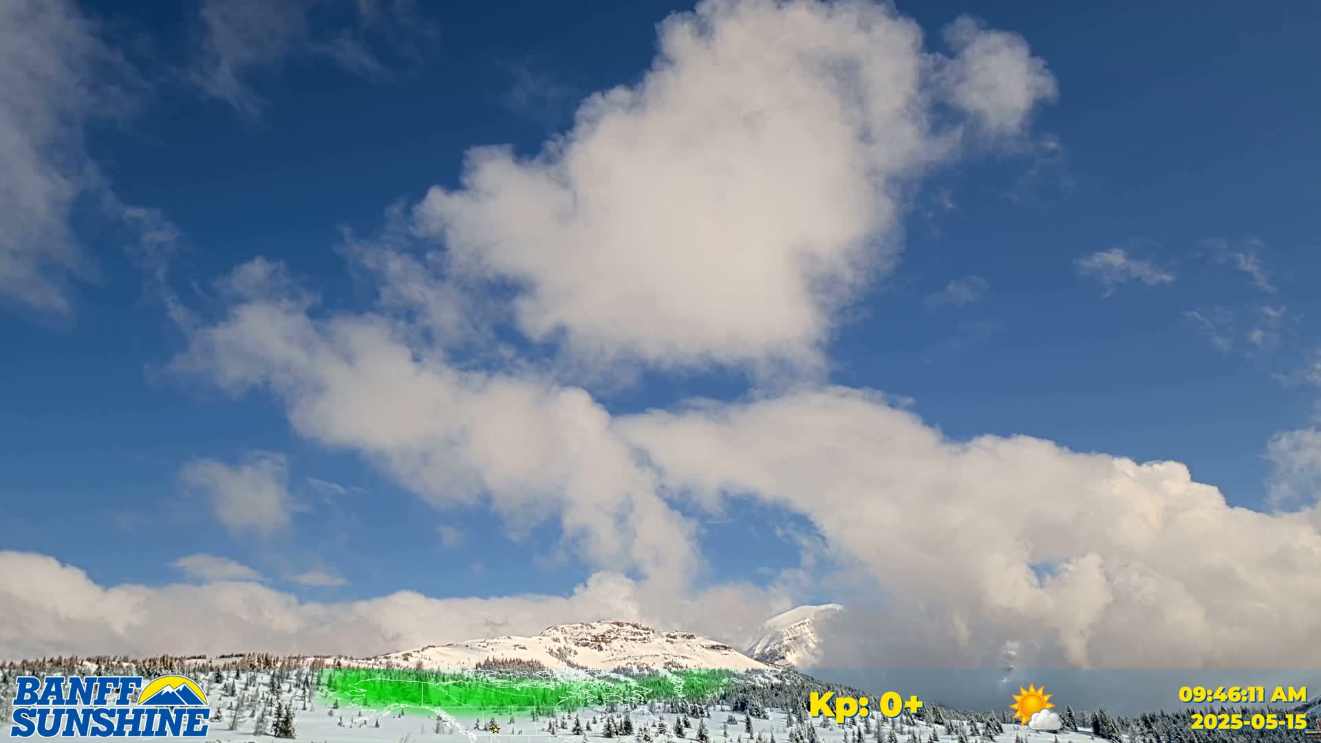 A partly cloudy, bright blue sky over a snow-covered mountain range with sparse, dark evergreen trees.