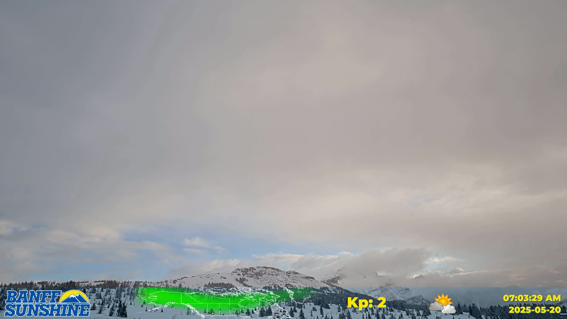 A mostly cloudy, pale sky hangs over snow-covered mountains and evergreen trees.