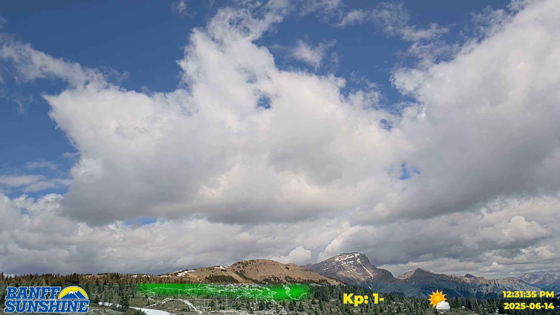 A mostly clear blue sky with scattered cumulus clouds overlooks a mountain range with some snow-capped peaks.