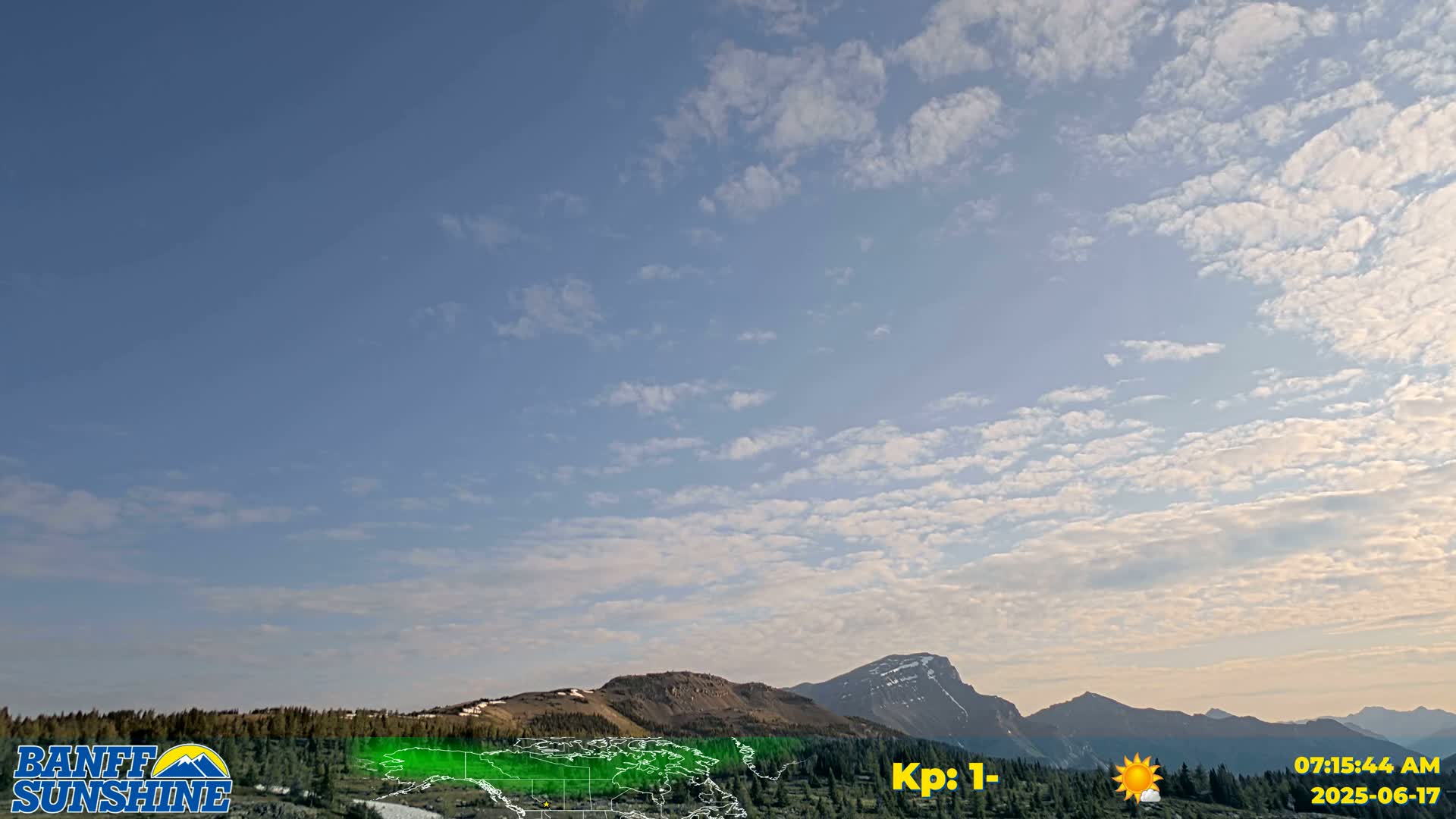 A mostly clear, blue sky with scattered clouds overlooks a mountain range and a valley of coniferous trees.