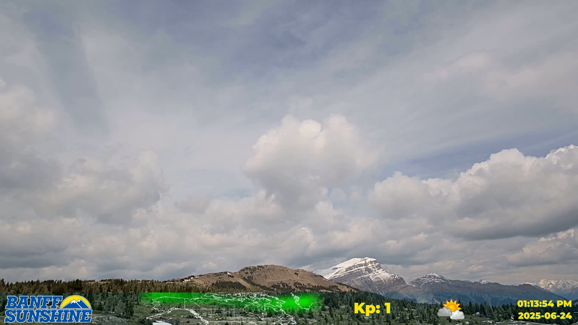 A partly cloudy sky overlooks a mountain range with patches of snow, and a foreground of dark green trees.