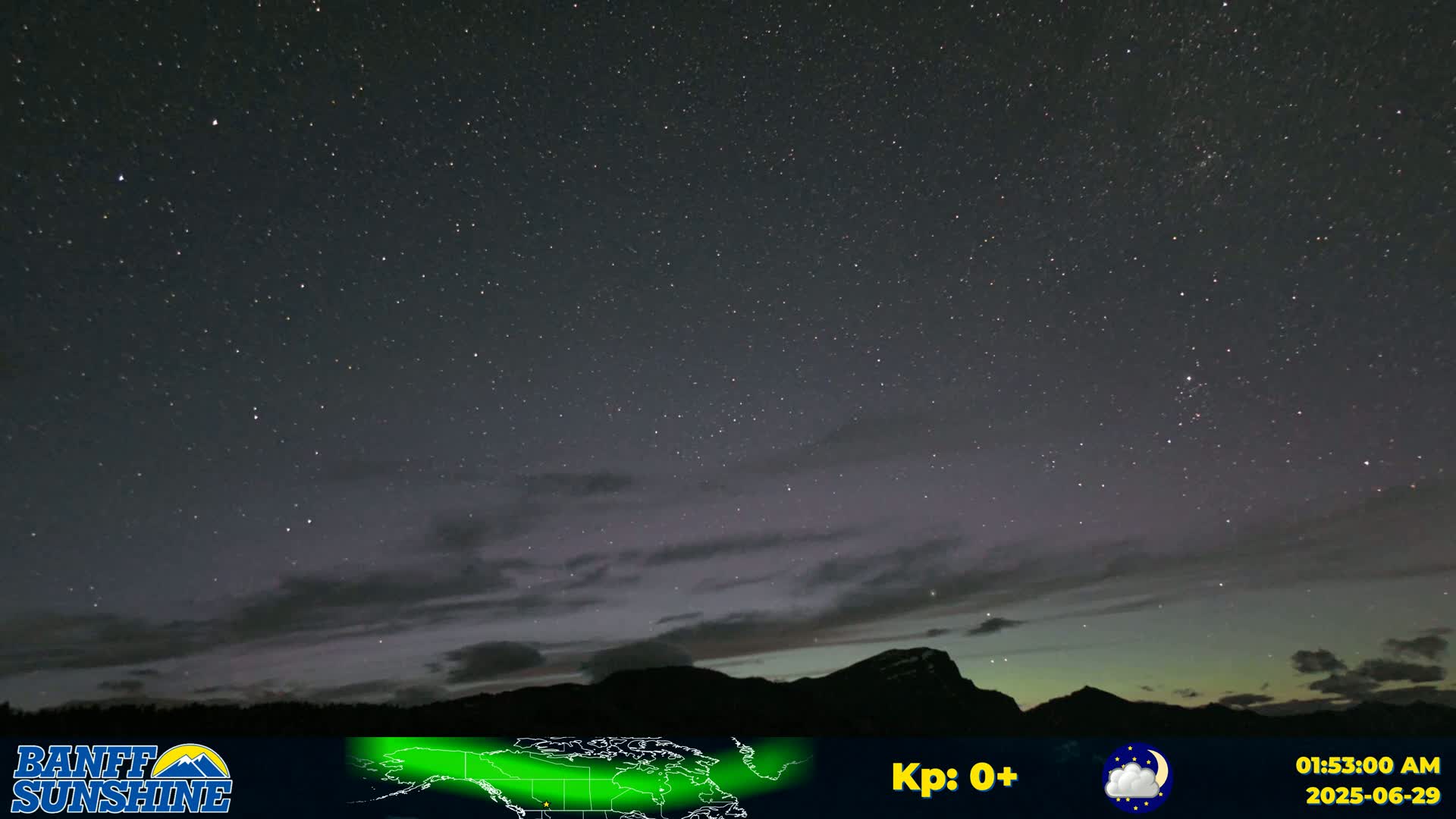 A nighttime view of a starry sky with a faint aurora borealis over a silhouetted mountain range under partly cloudy conditions.