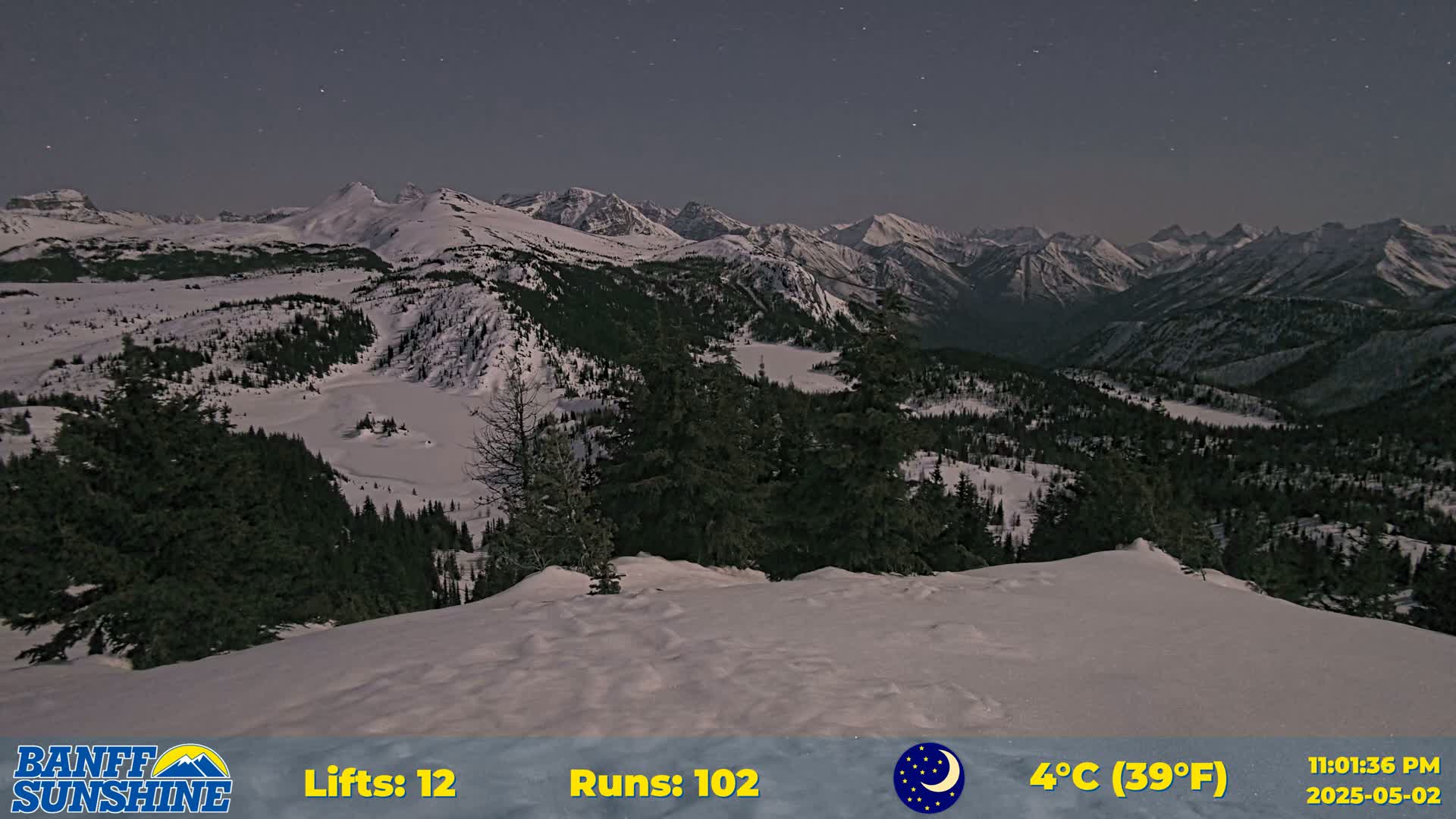 A snow-covered landscape at night, featuring a mountainous region with evergreen trees under a clear, starry sky, and a temperature of 4°C (39°F).