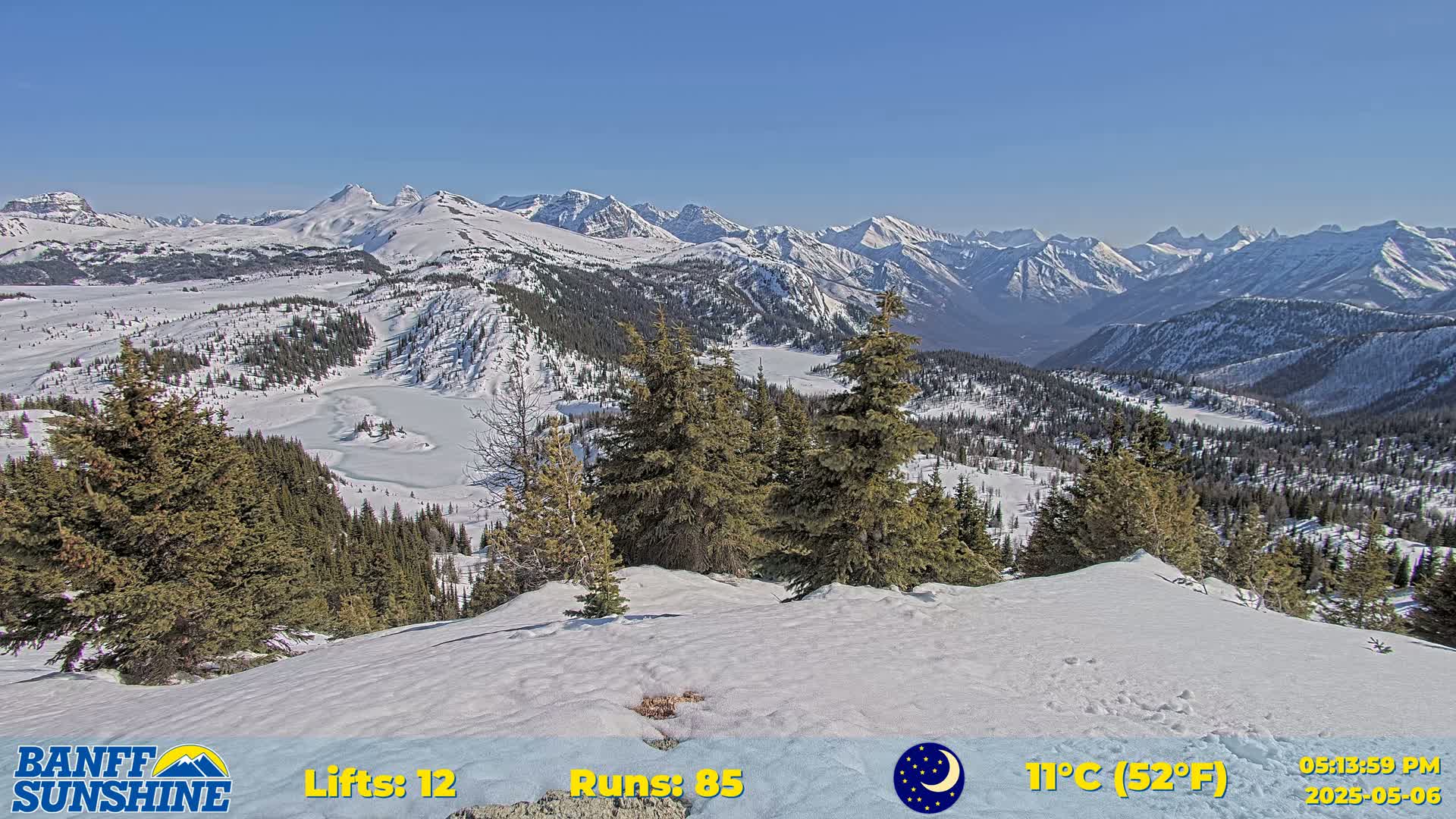 A snow-covered mountain vista reveals a partially frozen lake nestled among evergreen trees, under a clear blue sky.