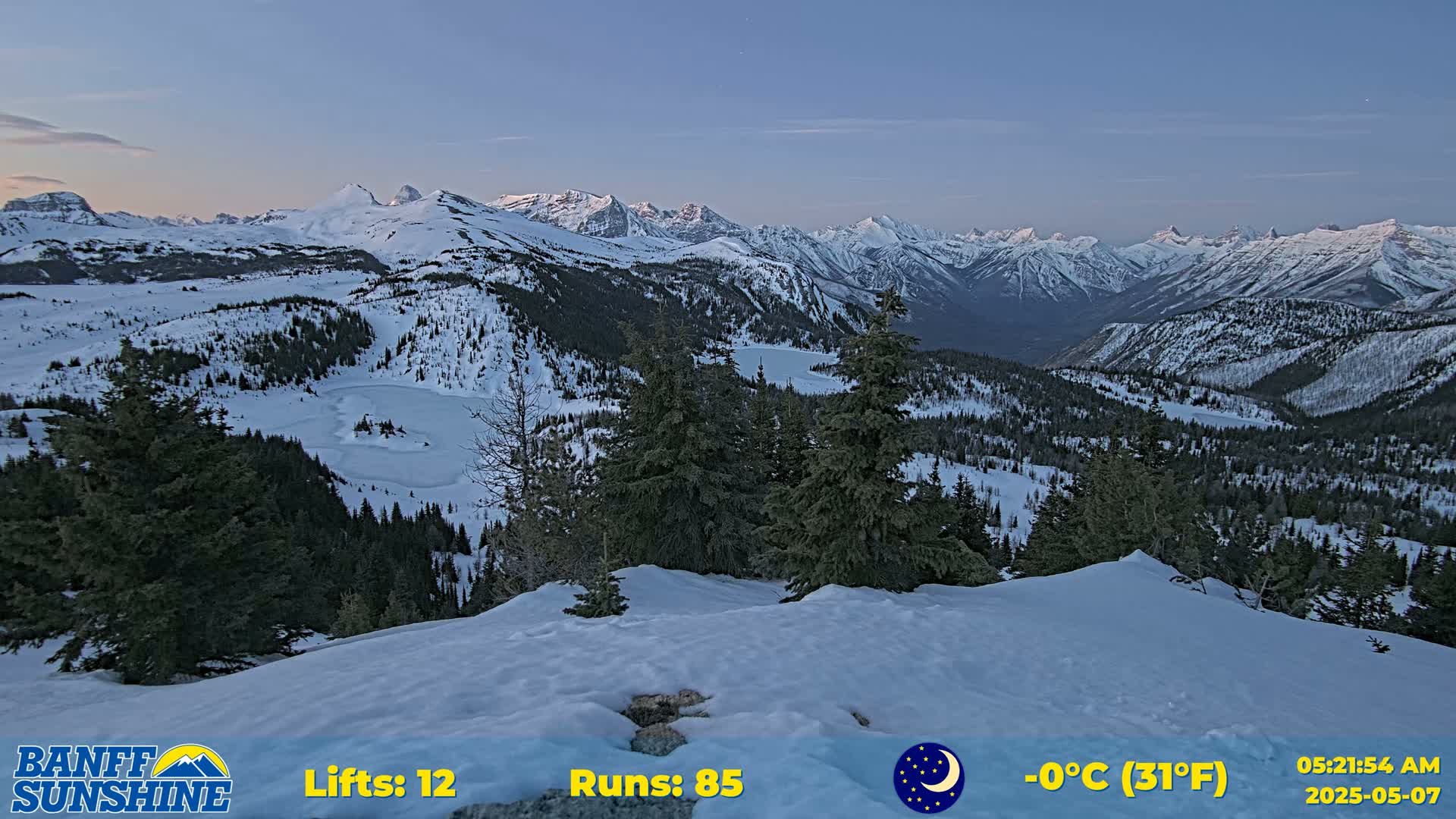 A snow-covered mountain range at twilight, with evergreen trees and a partially frozen lake visible in the valley below, at a temperature of 0°C (31°F).