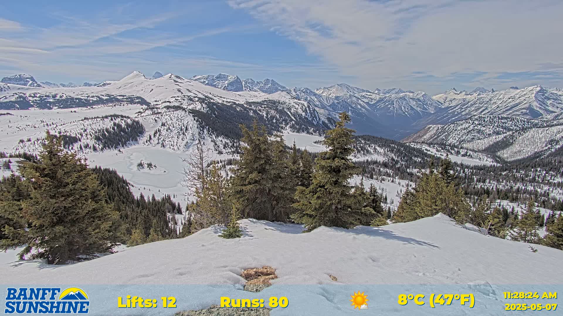 A snow-covered mountain range is viewed from a snowy peak under a partly sunny sky.