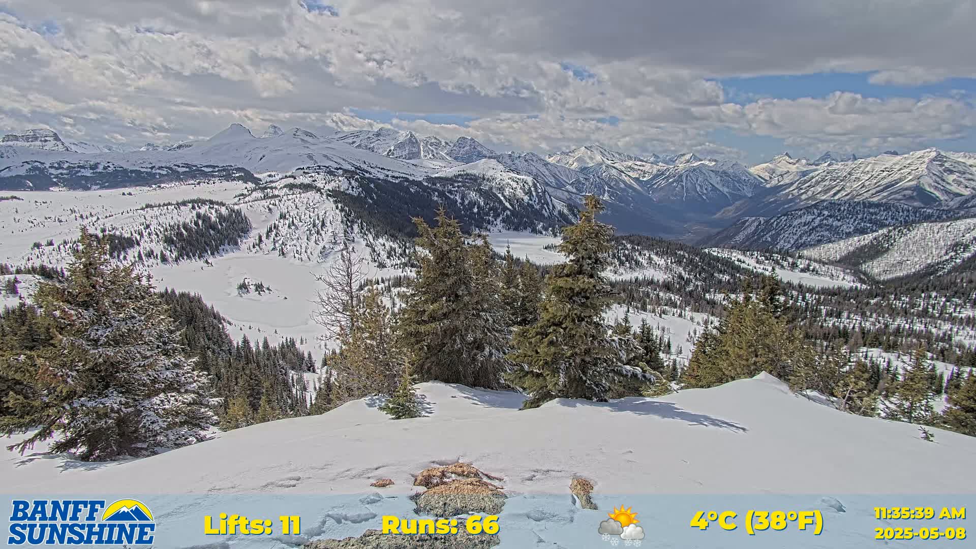 A snowy mountain range under a partly cloudy sky is viewed from a snow-covered peak.