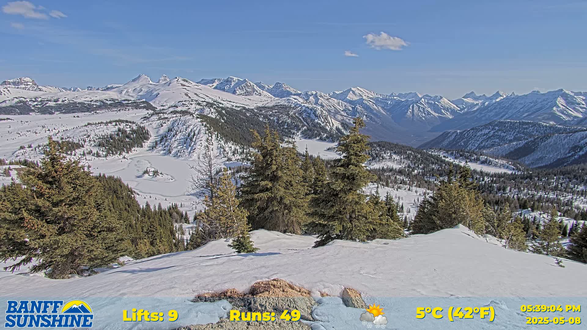 A snow-covered mountain range with a partly sunny sky and a partially frozen lake is visible from a snowy vantage point.
