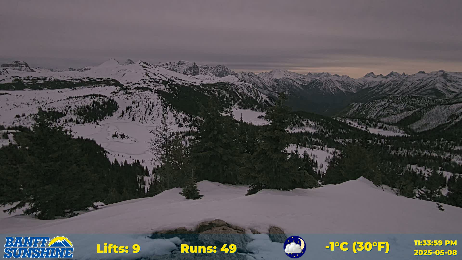A snow-covered mountain range is viewed from a snowy vantage point under a cloudy, dusky sky.
