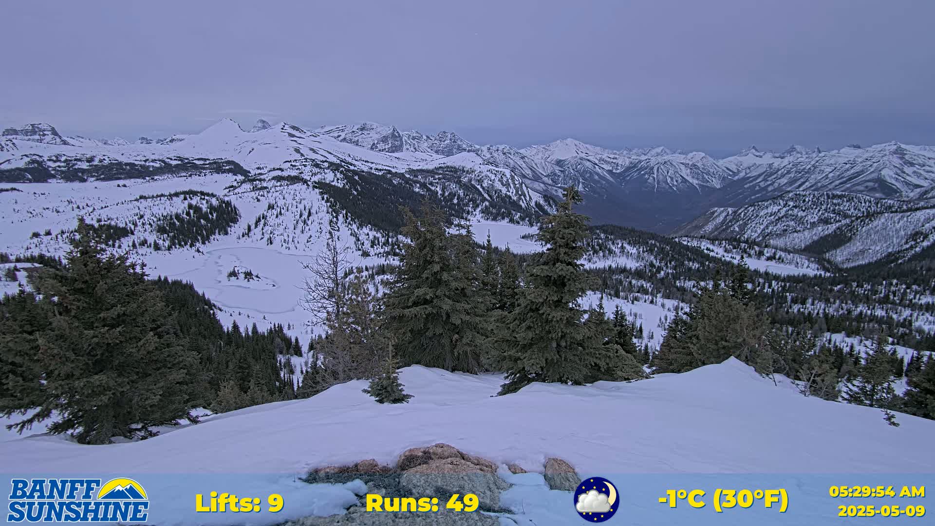 A snow-covered mountain landscape with evergreen trees under a twilight sky, showing a frozen lake in the valley below.