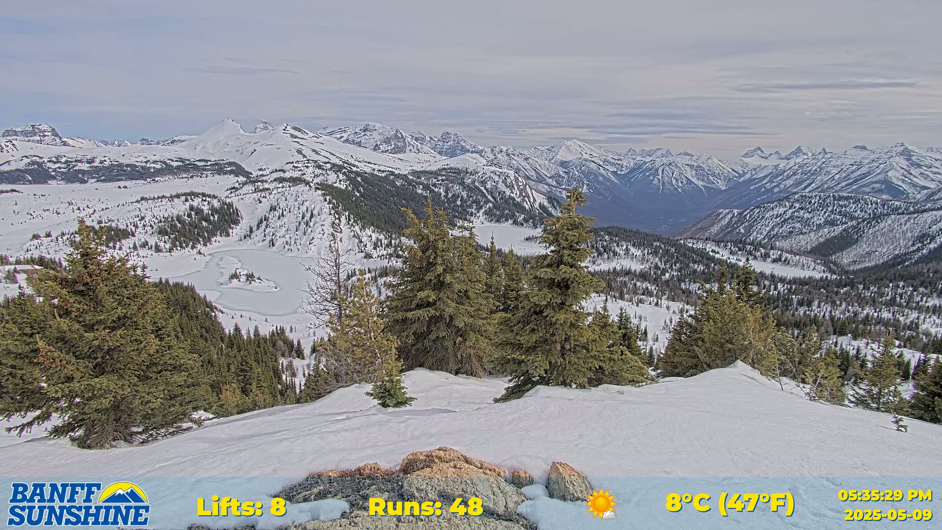A snow-covered mountain vista with evergreen trees in the foreground, under partly cloudy skies, shows a partially frozen lake in a valley below.