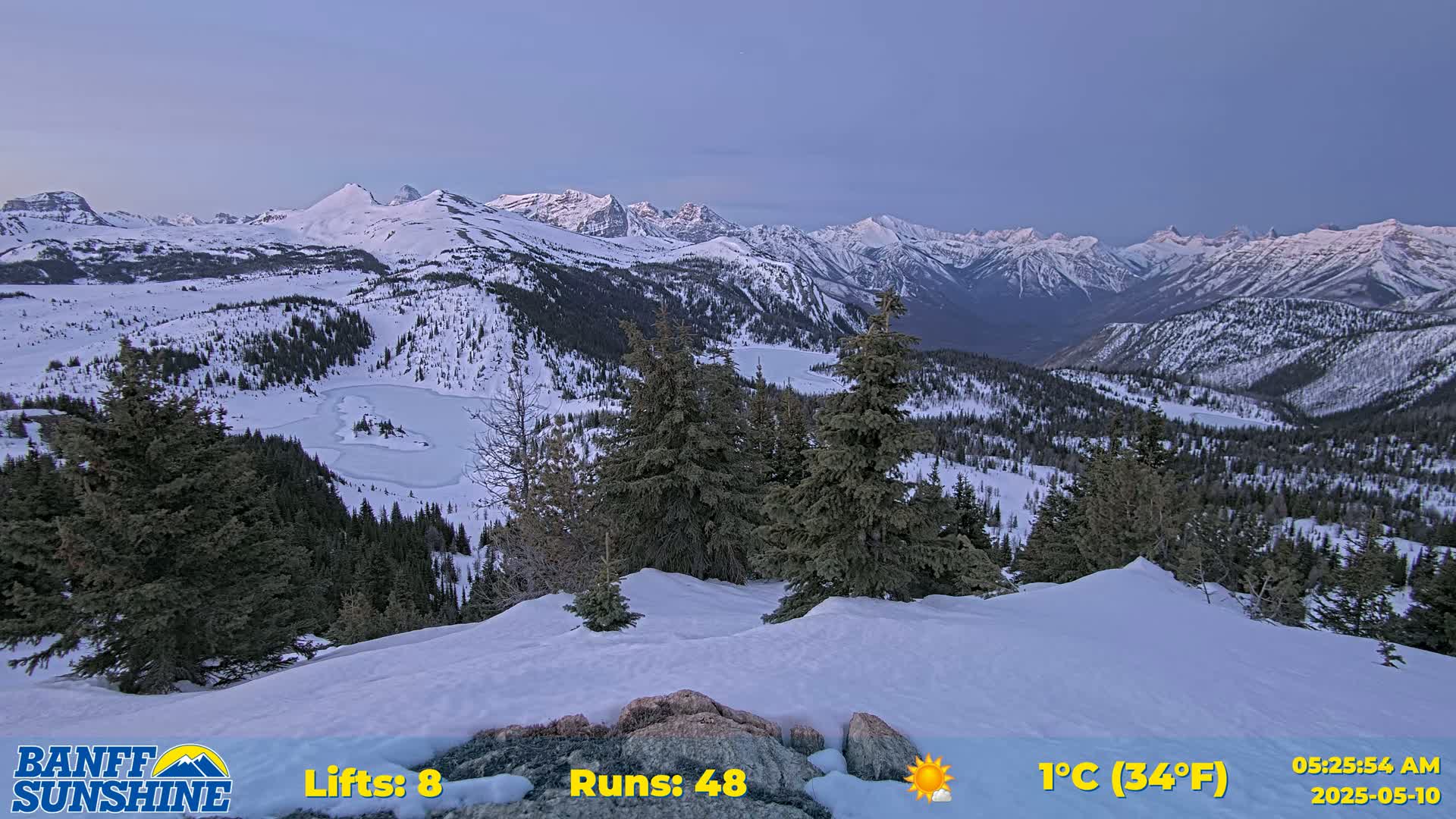A snow-covered mountain landscape at dawn, featuring a partially frozen lake nestled among evergreen trees and a range of snow-capped peaks in the background under a clear, pale blue sky.