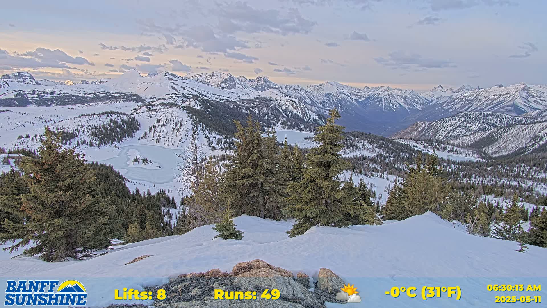 A snow-covered mountain landscape at sunrise, featuring a partially frozen lake, evergreen trees, and a mountainous backdrop under partly cloudy skies.