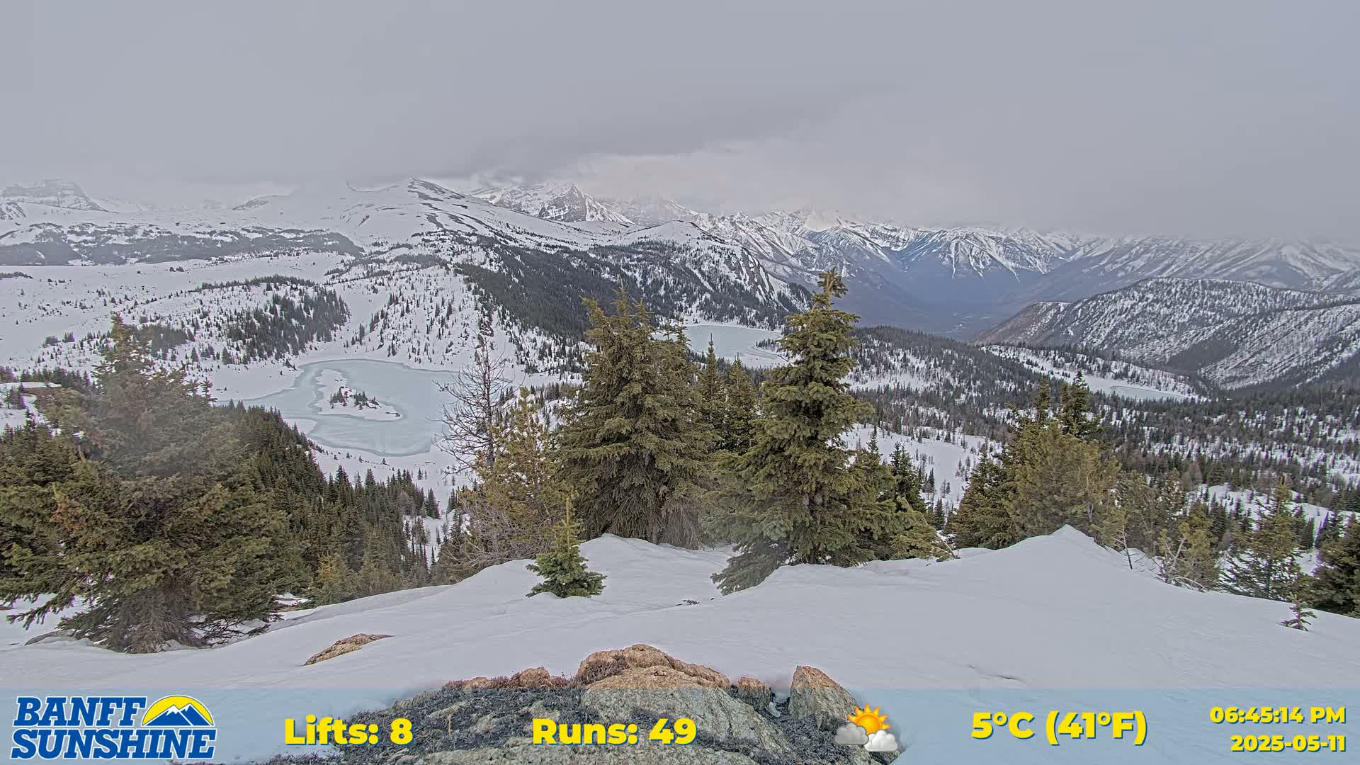 A snow-covered mountain vista with evergreen trees and partially frozen lakes is visible under an overcast sky.