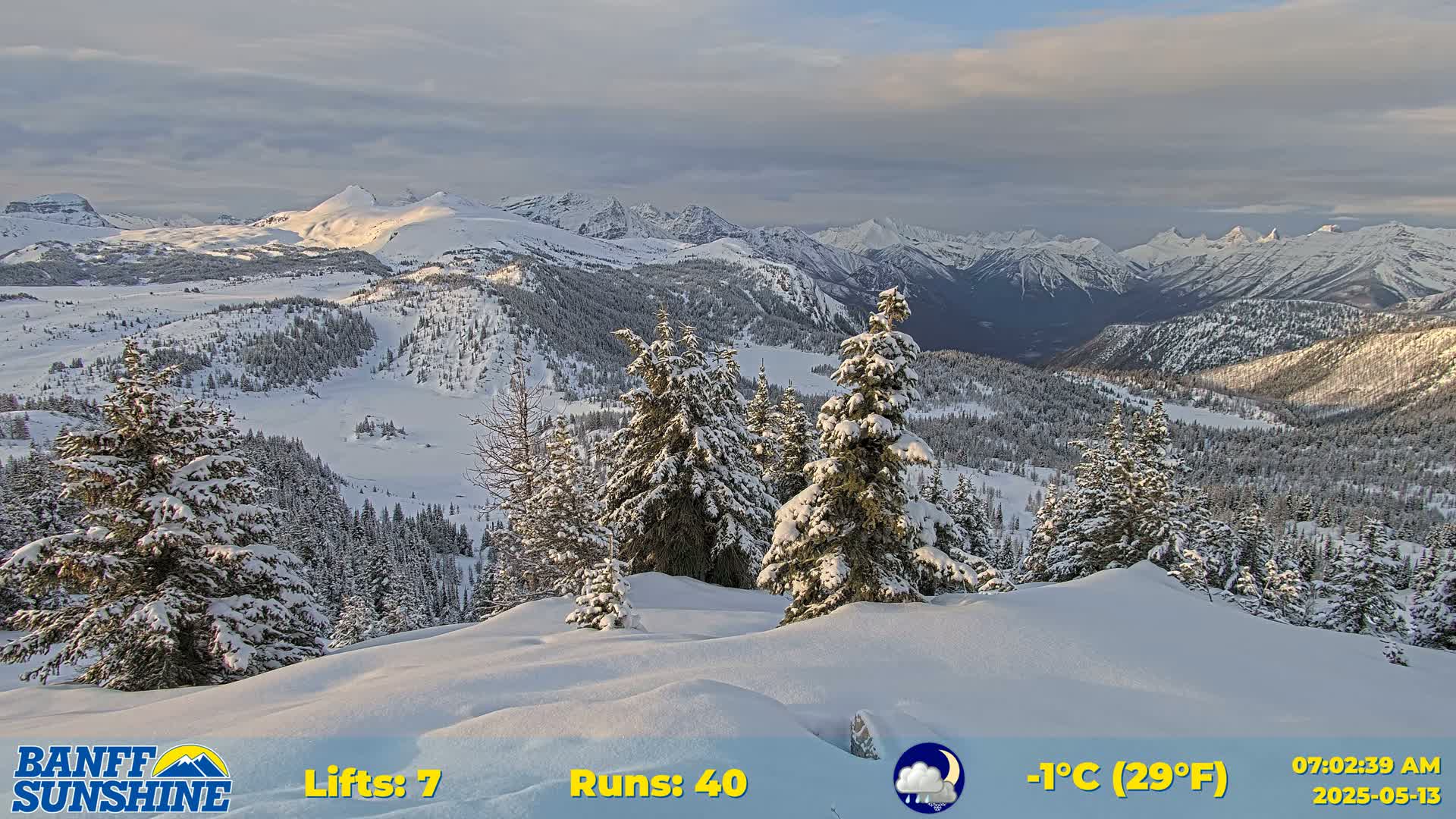 A snow-covered mountain range is viewed from a snowy vantage point with evergreen trees dusted with snow, under a partly cloudy sky.