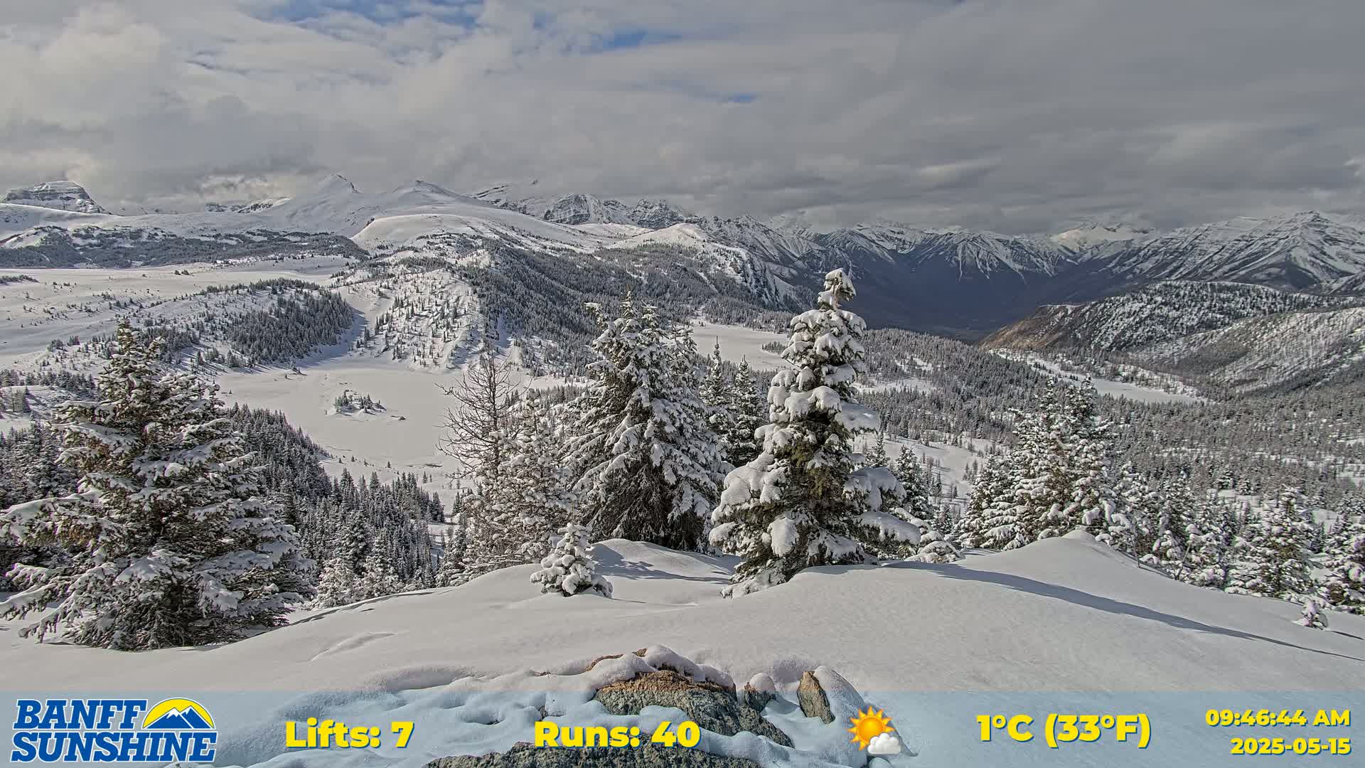 A snow-covered mountain landscape with partly sunny skies, featuring snow-laden evergreen trees in the foreground and a valley extending to distant snow-capped mountains in the background.