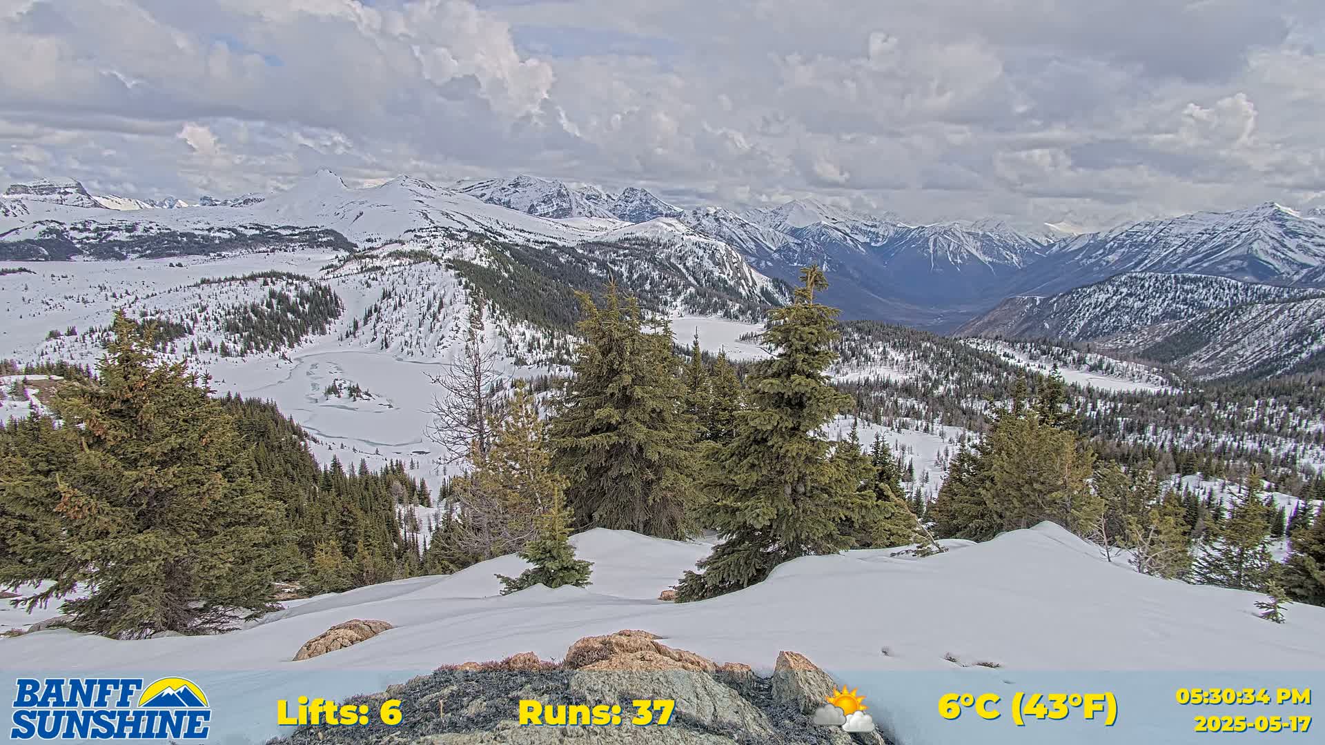 A partly cloudy, snowy mountain vista with evergreens and a partially frozen lake is visible from a snow-covered vantage point.